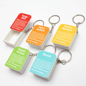 May include: Five small, white, plastic containers with keychains attached. Each container has a different colored label with a different flavor name: Tangy BBQ, Spicy Buffalo, Honey Mustard, Sweet 'N Sour, and Ranch. Each label includes a list of ingredients and a net weight of 1 oz (25.5g).