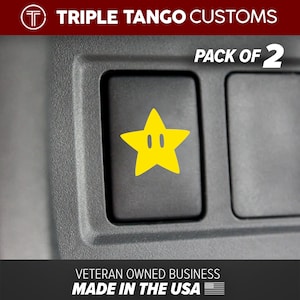May include: Two yellow star-shaped car decals with a black outline and two white circles for eyes. The decals are on a gray car button. The text "PACK OF 2" is above the buttons. The text "TRIPLE TANGO CUSTOMS" is at the top of the image. The text "VETERAN OWNED BUSINESS MADE IN THE USA" is at the bottom of the image.