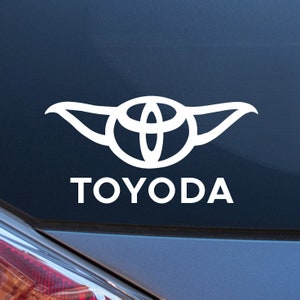 May include: A white vinyl sticker with a stylized Yoda-like figure with the Toyota logo in the center and the word "TOYODA" below it.