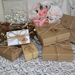 May include: A collection of small, brown gift boxes tied with twine and decorated with a small, red heart tag. The boxes are arranged on a white lace tablecloth with a vase of pink roses in the background.