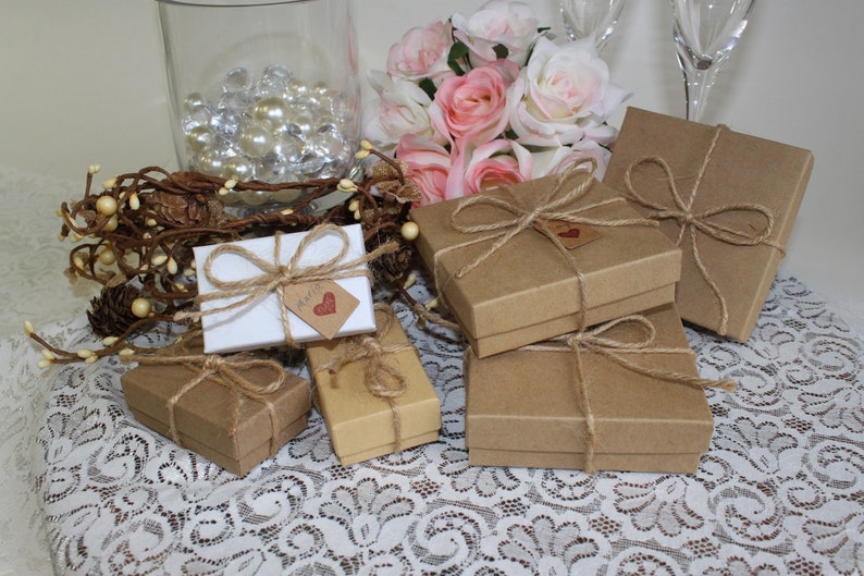 May include: A collection of small, brown gift boxes tied with twine and decorated with a small, red heart tag. The boxes are arranged on a white lace tablecloth with a vase of pink roses in the background.