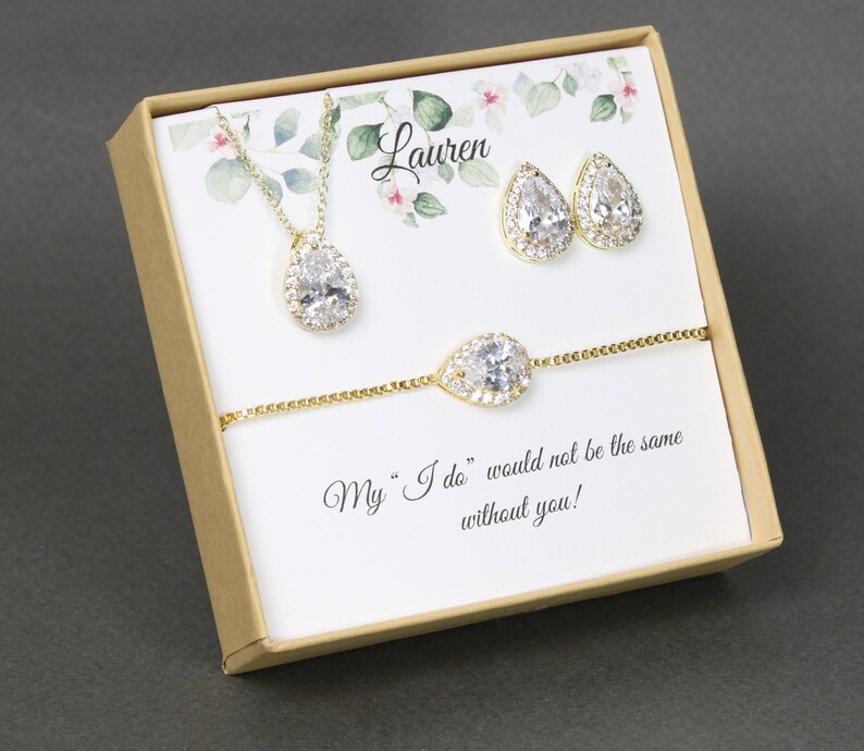 May include: A gift box with a gold necklace, earrings, and bracelet with pear-shaped clear stones. The box has a floral design and the text "Lauren" and "My "I do" would not be the same without you!"