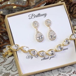 May include: A gold bridal jewelry set with a teardrop-shaped bracelet and matching earrings. The jewelry is in a box with the name "Brittany" and the text "My  without you!"