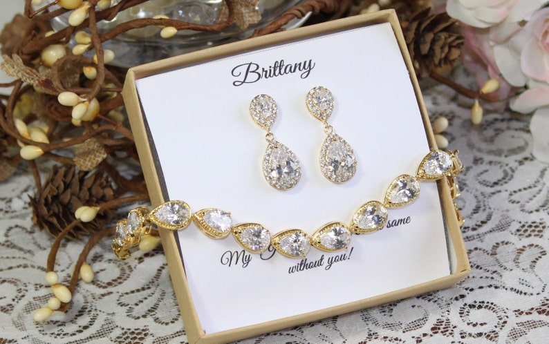 May include: A gold bridal jewelry set with a teardrop-shaped bracelet and matching earrings. The jewelry is in a box with the name "Brittany" and the text "My  without you!"