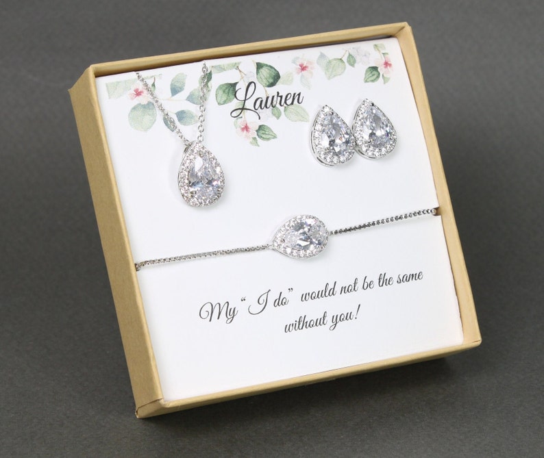 May include: A gift box with a personalized message for a bridesmaid. The box contains a silver necklace, earrings, and bracelet with teardrop-shaped crystals.