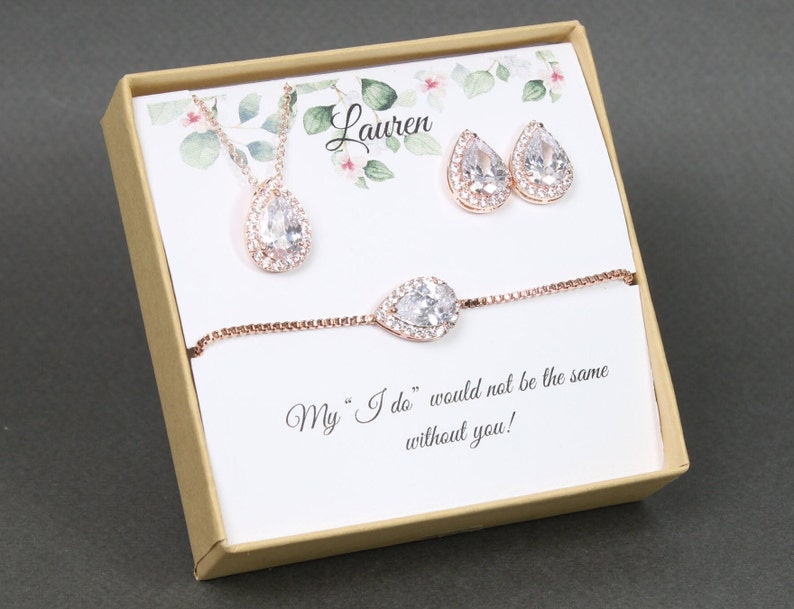 May include: A gift box with a rose gold necklace, earrings, and bracelet with pear-shaped clear stones. The box has a white card with the text "My "I do" would not be the same without you!" and the name "Lauren" printed on it.