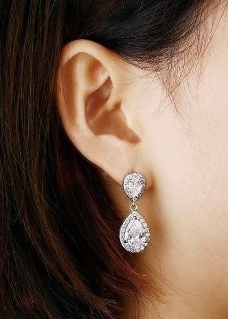 May include: A pair of silver teardrop earrings with clear cubic zirconia stones. The earrings have a double teardrop design, with a smaller teardrop stone on top of a larger teardrop stone.