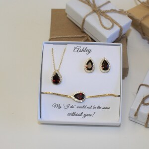 May include: A white gift box with a gold necklace, earrings, and bracelet with a red teardrop-shaped stone. The box has the name "Ashley" written on it and the text "My "I do" would not be the same without you!"