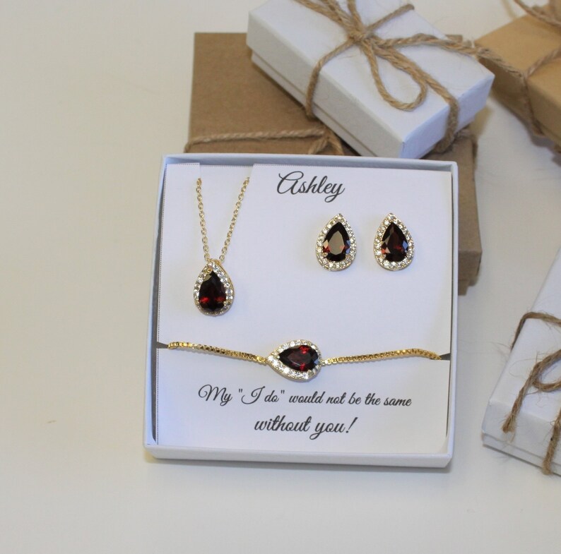May include: A white gift box with a gold necklace, earrings, and bracelet with a red teardrop-shaped stone. The box has the name "Ashley" written on it and the text "My "I do" would not be the same without you!"