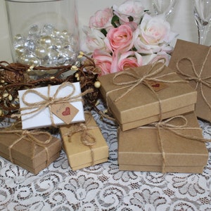 May include: A collection of small, brown gift boxes tied with twine and decorated with a small heart tag. The boxes are arranged on a white lace tablecloth with a vase of pink roses in the background.