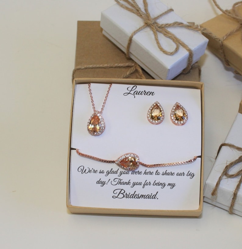 May include: A gift box with a rose gold necklace, earrings, and bracelet with a pear-shaped stone. The box has a personalized message that says "Lauren We're so glad you were here to share our big day! Thank you for being my Bridesmaid."