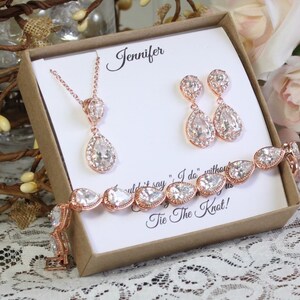 May include: A rose gold bridal jewelry set in a gift box. The set includes a necklace, earrings, and a bracelet. The jewelry features pear-shaped clear crystals with a rose gold setting. The box has a personalized message that says "Jennifer" and "Tie the Knot!"