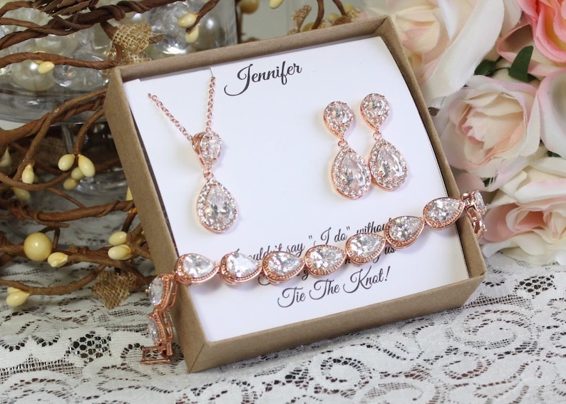 May include: A rose gold bridal jewelry set in a gift box. The set includes a necklace, earrings, and a bracelet. The jewelry features pear-shaped clear crystals with a rose gold setting. The box has a personalized message that says "Jennifer" and "Tie the Knot!"
