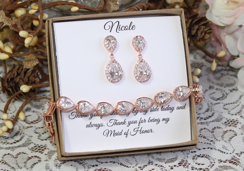 May include: A gift box with a white card that says "Nicole" and "Thank you for being my Maid of Honor." The box contains a rose gold bracelet and earrings with pear-shaped clear stones.
