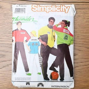 May include: A Simplicity sewing pattern envelope for Days of Thunder apparel, featuring illustrations of three individuals wearing racing-themed jackets and trousers in red, yellow, green, and white, with the number 51 and chequered flags visible.