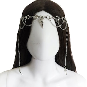 May include: Silver-toned headpiece featuring a central bird skull pendant and dangling chains. The headpiece is composed of interconnected rings and chains, with additional decorative elements. The design is symmetrical, with chains cascading down on either side.