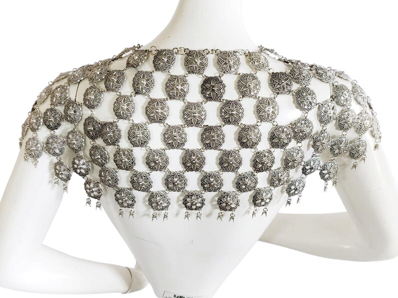 May include: Silver chainmail shoulder piece with a decorative design. The chainmail is made of small, round links that are connected to form a mesh-like fabric. The shoulder piece has a fringe at the bottom.