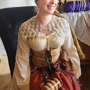 May include: A woman wearing a white blouse with a silver and white beaded collar, a brown corset, a brown skirt, and a brown leather belt. She is holding a wooden mug with a striped design. She is wearing a brown headscarf with a floral pattern.