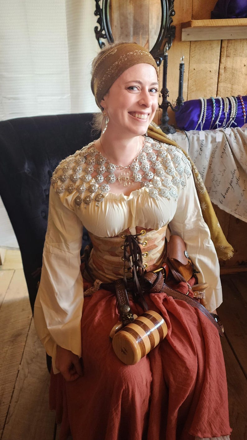 May include: A woman wearing a white blouse with a silver and white beaded collar, a brown corset, a brown skirt, and a brown leather belt. She is holding a wooden mug with a striped design. She is wearing a brown headscarf with a floral pattern.