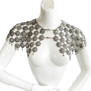 May include: A silver metal chainmail shoulder piece with a decorative floral pattern. The chainmail is made of small, interconnected metal rings.
