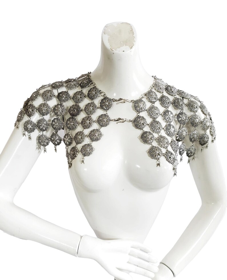 May include: A silver metal chainmail shoulder piece with a decorative floral pattern. The chainmail is made of small, interconnected metal rings.