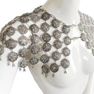 May include: Silver chainmail shoulder piece with a floral design. The chainmail is made of small, interconnected metal rings. The piece is designed to be worn over one shoulder and drapes down the chest.