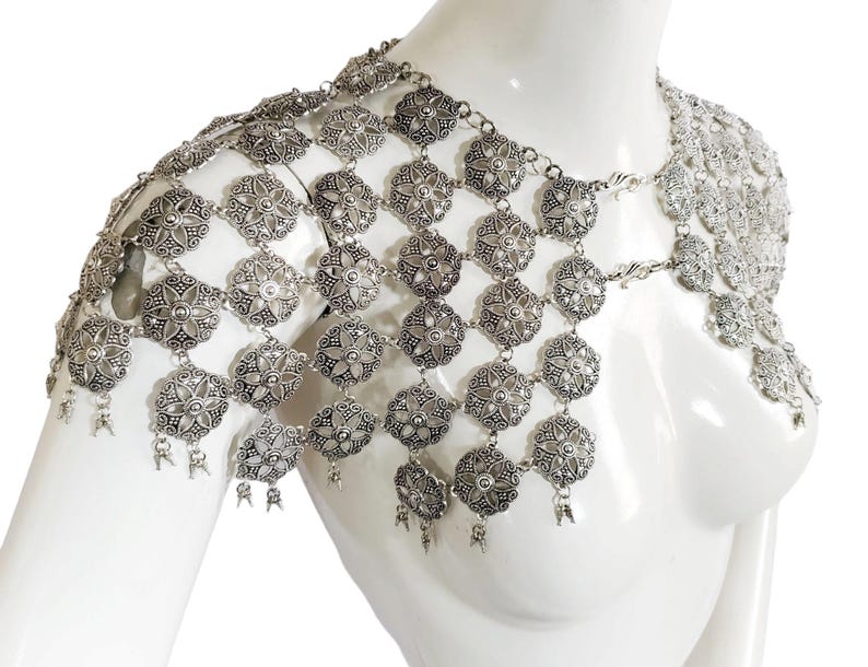 May include: Silver chainmail shoulder piece with a floral design. The chainmail is made of small, interconnected metal rings. The piece is designed to be worn over one shoulder and drapes down the chest.