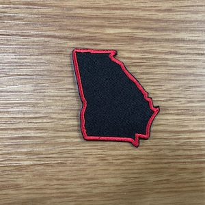 May include: A black patch in the shape of the state of Georgia with a red border. The patch is on a wooden surface.