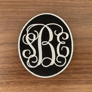 May include: A circular embroidered patch featuring a black background with a white border. The center displays a large, white, ornate, swirling monogram of the letters S, B, and E, stitched in a decorative, cursive-style font.