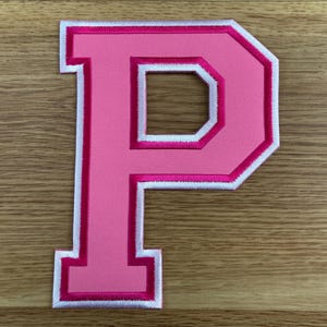 May include: A pink embroidered letter "P" patch with a white border. The letter has a block letter style with a white inner border. The patch is on a wooden surface.