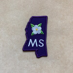 May include: A purple embroidered patch in the shape of the state of Mississippi. The patch features a white flower with green leaves and the letters "MS" in white. The patch has a purple border.