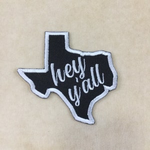 May include: Black embroidered patch in the shape of Texas with a white border. The phrase "hey y'all" is stitched in white cursive across the center of the patch. The patch is on a beige background.