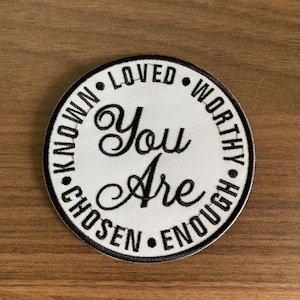 May include: A round, white patch with black text. The words "You Are" are in the center, with "LOVED", "WORTHY", "ENOUGH", "CHOSEN", and "KNOWN" circling the edge. The patch has a black border and is set against a wood grain background.