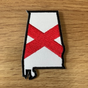 May include: An embroidered patch in the shape of the state of Alabama. The patch features a white background with a bold red saltire cross, all outlined with a thick black border. The embroidery has a visible diagonal texture throughout the design.