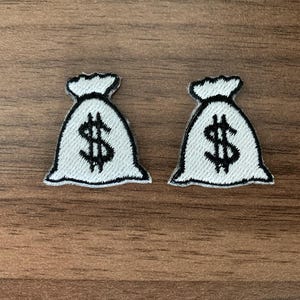 May include: Two embroidered patches, each shaped like a white money bag with a black outline and a black dollar sign symbol centered on the front. The patches feature a tied top section and a rounded base, rendered in a clean, high-contrast design.