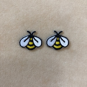 May include: Two embroidered bee patches. Each patch features a white wing design with black outlines and a yellow and black striped body. The patches are small and detailed, perfect for adding to clothing or accessories.