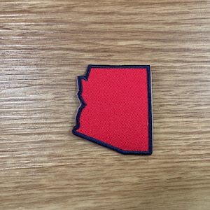 May include: A red patch in the shape of the state of Arizona, with a navy blue border. The patch is on a wooden surface.