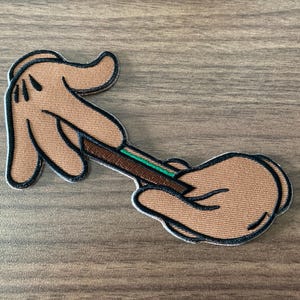 May include: Embroidered patch featuring cartoon hands holding a rolled cigarette. The hands are tan with black outlines and details. The cigarette is brown with green and white accents. The patch is on a wooden surface.