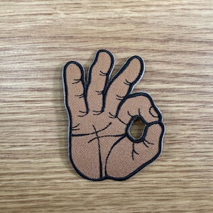May include: A brown patch in the shape of a hand making the "OK" gesture. The patch has a black border and is set against a wooden background. The fingers are slightly curved, and the thumb and index finger form a circle.