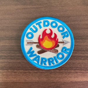 May include: A round patch with the words "OUTDOOR WARRIOR" in light blue, surrounding a campfire graphic. The campfire has red and yellow flames, with two arrows crossing behind it. The patch is on a wooden surface.