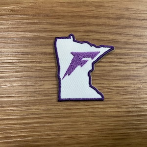 May include: A white patch in the shape of the state of Minnesota, with a purple outline and a purple stylized letter "F" inside. The patch is on a wooden surface.