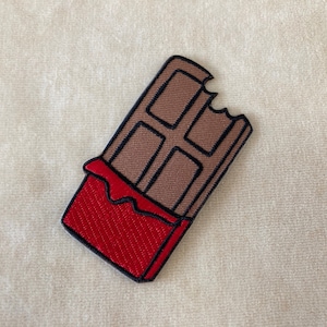 May include: Embroidered patch of a chocolate bar with a bite taken out. The chocolate is brown with black outlines, and the wrapper is red. The patch is on a cream-colored fabric.