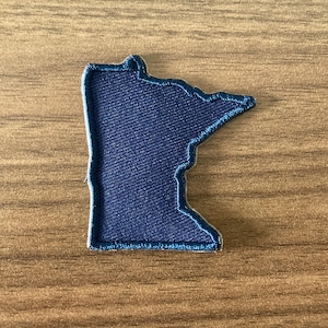 May include: A dark navy blue embroidered patch in the shape of the state of Minnesota. The patch features a dense, textured thread construction with a clean, stitched border that follows the distinct geographic outline of the state.