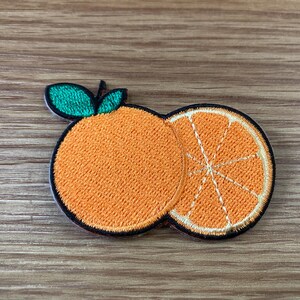 May include: Embroidered orange patch featuring a whole orange and a sliced orange half. The patch has a black border and green leaves. The orange is a vibrant orange color, and the background is a wooden surface.