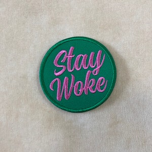 May include: A round, green embroidered patch with the words "Stay Woke" in pink script. The patch has a green border and is set against a neutral background.