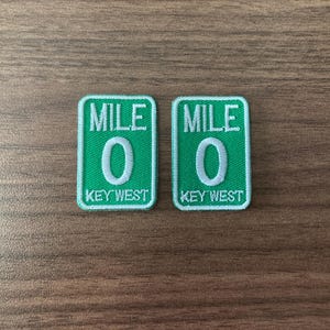 May include: Two green embroidered patches with white text and a white border. Each patch reads "MILE 0" above "KEY WEST". The patches are rectangular with rounded corners.