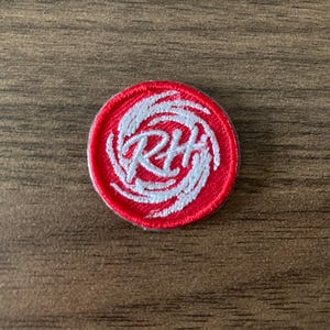 May include: A round, red embroidered patch with white swirling designs and the letters "RH" in the center. The patch is circular and has a white border. The patch is on a wooden surface.
