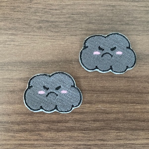 May include: Two embroidered patches shaped like dark gray clouds with a grumpy facial expression, featuring stitched black eyes and mouths, along with small pink blush marks on the cheeks, outlined with a thin white border for clear definition.