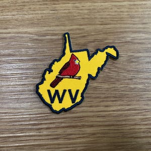 May include: Embroidered patch in the shape of West Virginia, featuring a red cardinal perched on a branch. The patch is yellow with a dark blue border and the letters "WV" in dark blue.