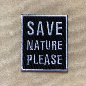 May include: Square black patch with white embroidered text that reads "SAVE NATURE PLEASE." The patch has a white border and is set against a beige background. The patch is likely an accessory.
